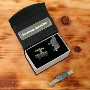 Peut inclure: Une boîte cadeau noire avec une plaque argentée indiquant "Your Message Engraved Here". La boîte contient deux boutons de manchette en argent et une pince à cravate, tous ornés d'un motif de mouton.