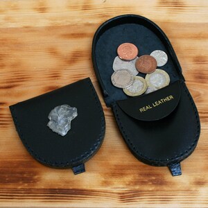 May include: Two black leather coin purses, one open and filled with coins. The open purse has "REAL LEATHER" printed on the flap. The other purse has a silver dog head emblem. Both are on a wooden surface.