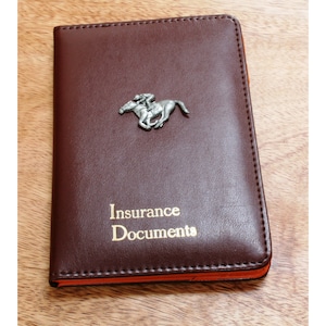 Puede incluir: Un portadocumentos de seguro de cuero marrón con una figura de plata de un caballo y un jinete en la parte delantera. El portadocumentos tiene las palabras "Insurance Documents" impresas en oro en la parte delantera.