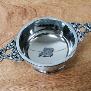 May include: A silver Celtic knot design handled cup with a shiny, reflective interior. Two small, silver, Celtic knot designs are inside the cup.