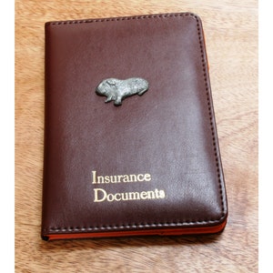 May include: A brown leather insurance document holder with a small silver guinea pig figurine on the front. The holder has the words "Insurance Documents" embossed in gold lettering.