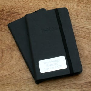May include: Two black notebooks with elastic closures. The top notebook has the word "notes" embossed on the front cover. A silver label on the front cover reads "Your Message Engraved Here For FREE".
