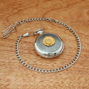 May include: Silver pocket watch with a gold sunflower design on the face, attached to a silver chain. The watch has a textured gray background and a silver casing. The chain is draped around the watch on a wooden surface.