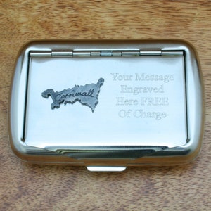 May include: A silver metal cigarette case with a hinged lid. The case has a small metal cutout of the shape of Cornwall, England, with the word "Cornwall" inscribed on it. The case also has the text "Your Message Engraved Here FREE Of Charge" engraved on it.