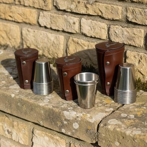 Könnte beinhalten: Ein Set aus Edelstahl-Schnapsgläsern mit braunen Leder-Tragetaschen. Die Schnapsgläser haben ein geriffeltes Design und werden auf einer Steinoberfläche präsentiert. Die Lederetuis haben einen Knopfverschluss.