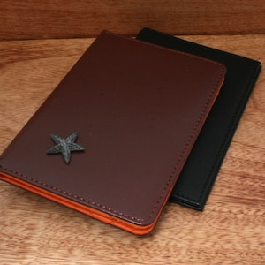 May include: A brown leather notebook with an orange interior and a small silver star design. The notebook is closed and sitting on a wooden surface.