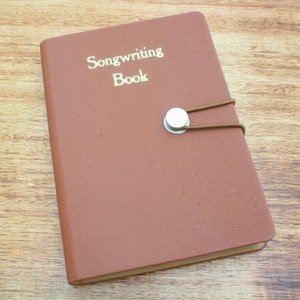 May include: A brown leather-bound songwriting book with a silver button closure and a brown elastic band. The book has the words "Songwriting Book" embossed in gold on the front cover.