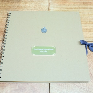 May include: A brown spiral bound scrapbook with a silver metal embellishment and a green label that says "Your Message Engraved Here For Free". A blue ribbon is tied around the book.