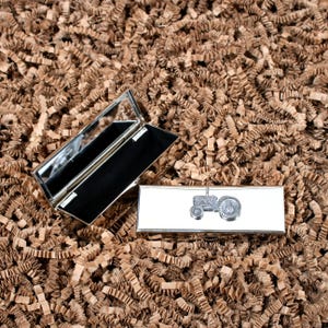 May include: A silver compact mirror with a black interior and a small tractor design on the front. The mirror is open and sitting on a brown paper surface.