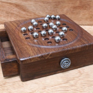 Könnte beinhalten: Ein Holzspielbrett mit einer Schublade und einer Metalleinlage. Das Brett hat eine kreisförmige Vertiefung mit 30 Löchern und 30 silbernen Metallkugeln.