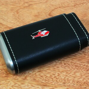 Puede incluir: Una funda de cuero negro con ribete plateado y un diseño de helicóptero rojo. La funda tiene costuras blancas alrededor de los bordes.