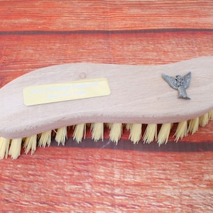 Può includere: Uno spazzolino di legno con manico curvo e setole gialle. Il manico ha una placca dorata con il testo "Your Name Engraved Here" e un piccolo ciondolo a forma di angelo d'argento.