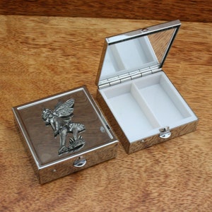 Puede incluir: Una caja de pastillas de plata con una tapa con bisagras y un espejo en el interior. La caja tiene un diseño en relieve de un hada en un hongo.
