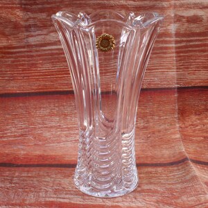 May include: A clear crystal vase with a flared, scalloped rim. The vase has vertical, curved ridges and a small, gold-toned emblem. The vase is designed for flowers and is approximately 25 cm tall.