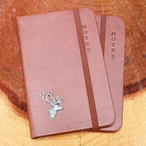 May include: Two brown leather bound notebooks with the word "NOTES" embossed on the front. The notebook on the left has a silver stag head design.