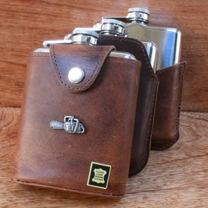 May include: Two stainless steel hip flasks with a brown leather carrying case. The case has a metal chainsaw charm attached to it. The case has a label that reads "Echtes Leder Leather".