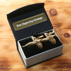 Könnte beinhalten: Goldfarbene Manschettenknöpfe in einer schwarzen und grauen Geschenkbox. Die Manschettenknöpfe zeigen ein Design mit zwei fliegenden Fasanen. Die Box hat ein goldenes Etikett mit der Aufschrift "Kostenlose Gravur Nachricht".