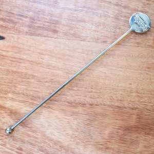 Könnte beinhalten: Ein silberner Rührstab aus Metall mit einem runden, flachen Ende. Das Ende hat ein dekoratives Design aus einer Reihe von Kreisen.