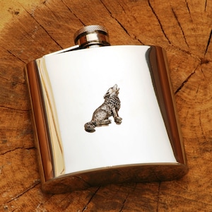 Op de afbeelding: Een gepolijste zilveren heupfles met een gedetailleerd huilende wolf-embleem. De fles heeft een afgeronde rechthoekige vorm en een schroefdop. De wolf is afgebeeld in een gedetailleerde, driedimensionale stijl.