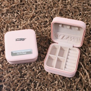 Könnte beinhalten: Zwei rosa, quadratische Schmuckkästchen. Eines ist geöffnet und zeigt Fächer zur Aufbewahrung. Das andere ist geschlossen, mit dem Text "Your Message Engraved Here Free Of Charge". Beide Boxen stehen auf einer braunen, strukturierten Oberfläche.