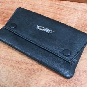 Könnte beinhalten: Eine schwarze Tabaktasche aus Leder mit einem silbernen Metallakzent. Die Tasche hat einen Klappverschluss und zwei Druckknöpfe.