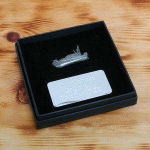 Puede incluir: Un pin plateado con forma de barco, presentado en una caja negra con interior de terciopelo negro. La caja también contiene una placa rectangular plateada con texto grabado. El pin es una miniatura detallada de un barco.