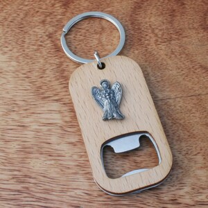 May include: A wooden bottle opener keychain with a silver angel charm. The keychain is made of light brown wood and has a silver metal bottle opener.