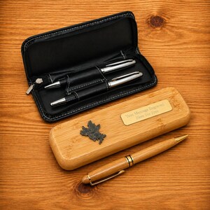 May include: A wooden pen set with a bamboo pen case and a black leather pen case. The bamboo case has a metal plate that reads "Your Message Engraved Here For Free". The set includes two silver pens and a wooden pen.