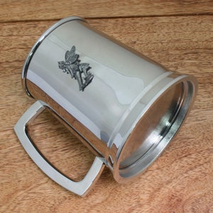 Puede incluir: Un tanque de metal plateado con un diseño de hada en la parte delantera. El tanque tiene un asa y una tapa.