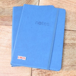 May include: Two blue notebooks with a black elastic band closure. The top notebook has the word "notes" embossed on the front cover. The bottom notebook has a red and white sticker with the word "LOVE" on it.