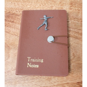 May include: Brown leather training notebook with a silver figure skater charm and a gold inscription that reads "Training Notes".