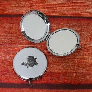 May include: A silver compact mirror with a design of a combine harvester on the front. The mirror is open and the inside is reflective.