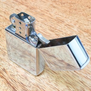 May include: A silver lighter with a hinged lid open, revealing the flint and wick. The lighter has a textured surface and is sitting on a wooden surface.