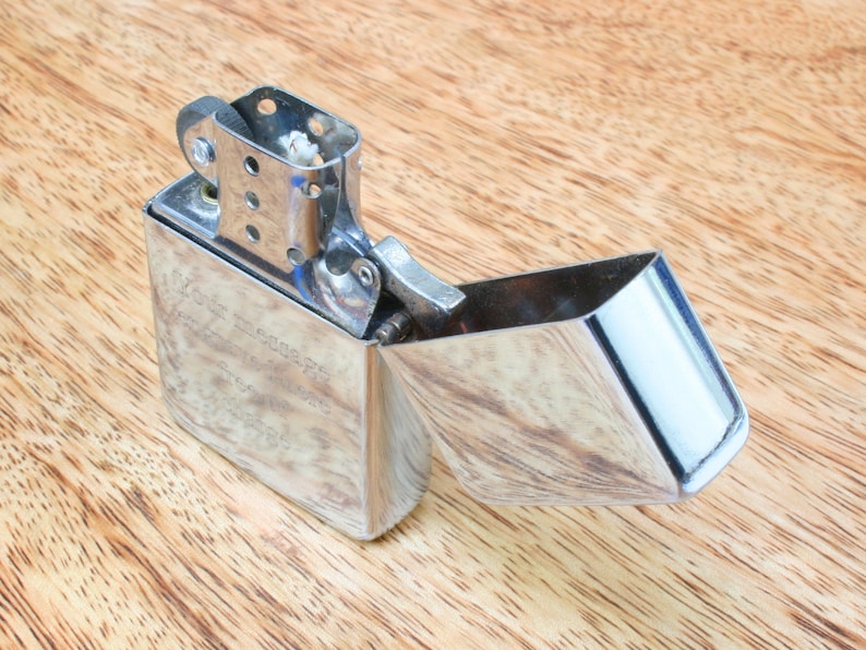 May include: A silver lighter with a hinged lid open, revealing the flint and wick. The lighter has a textured surface and is sitting on a wooden surface.