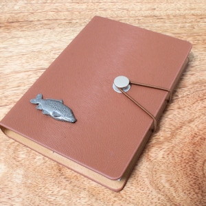 May include: A brown leather journal with a silver fish charm and a brown leather strap with a silver button closure.