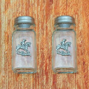 Op de afbeelding: Twee glazen zout- en peperstrooiers van helder glas met zilveren deksels. Elke strooier heeft een tinnen ontwerp van een ridder te paard die een draak doodt.