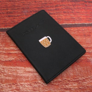 May include: Black leather passport holder with the word "PASSPORT" embossed on the front. A small gold metal beer mug pin is attached to the front.