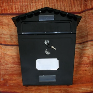 Puede incluir: Buzón de metal negro con una ranura para el correo, una cerradura y una placa de nombre en blanco. El buzón tiene una parte superior festoneada decorativa.