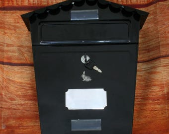 Può includere: Cassetta postale in metallo nero con una fessura per la posta, un buco della serratura e una targhetta vuota. La cassetta postale ha una parte superiore decorata a festoni.