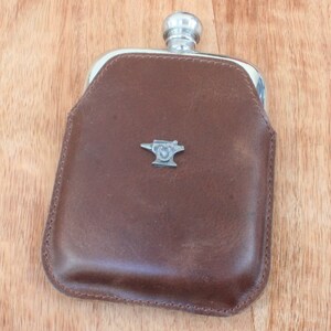 Puede incluir: Una petaca de cuero marrón con tapa de metal plateado y un emblema de yunque de metal plateado.