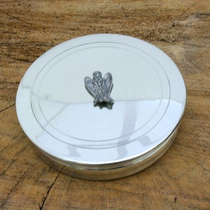 Puede incluir: Una caja redonda de plata para alhajas con un diseño de ángel en relieve en la tapa.