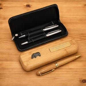 K&ouml;nnte beinhalten: Ein schwarzes Leder-Stiftetui mit zwei silbernen Stiften und ein Stiftetui aus Holz mit einem Nilpferd-Emblem. Das Holz-Etui hat eine goldene Plakette mit der Aufschrift "Your Message Engraved Here For Free". Ein Holzstift mit goldenen Akzenten ist ebenfalls vorhanden.