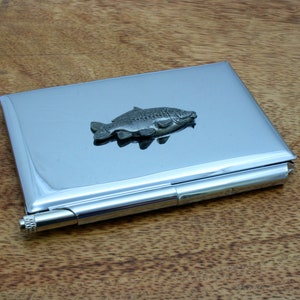 Puede incluir: Un porta tarjetas de metal plateado con un diseño de pez carpa 3D. El porta tarjetas tiene un bolígrafo incorporado.