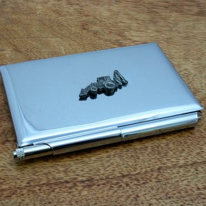 May include: A silver metal business card holder with a small tractor design on the front. The holder has a built-in pen.