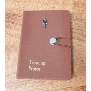 May include: A brown leather notebook with a gold inscription that reads "Training Notes". A small silver button and a black silhouette of a person are attached to the front cover.