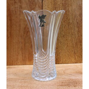 May include: A clear glass vase with a decorative, textured design. The vase has a wide mouth and a narrow base. A silver fairy figurine is attached to the side of the vase.