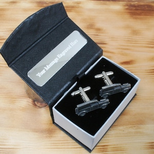 Puede incluir: Una caja de gemelos negra con una placa plateada que dice "Your Message Engraved Here". La caja contiene dos gemelos plateados con un diseño de coche negro.