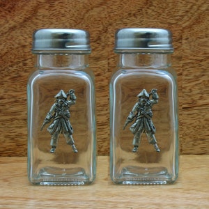 Może przedstawiać: Zestaw dwóch szklanych solniczek i pieprzniczek ze srebrnymi pokrywkami. Każda solniczka zawiera ołowianą figurkę pirata trzymającego miecz i butelkę.
