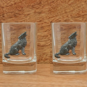 Puede incluir: Dos vasos de chupito de vidrio transparente con una figurita de lobo de peltre aullando a la luna. El lobo está sentado en el fondo del vaso.