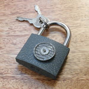 Puede incluir: Un candado gris con una cerradura plateada y un diseño de escudo decorativo en la parte delantera. El candado está sobre una superficie de madera y tiene dos llaves sujetas a él.